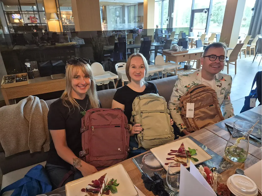 Three colleagues at a restaurant table, each holding a new travel backpack, plates and glasses in front of them.