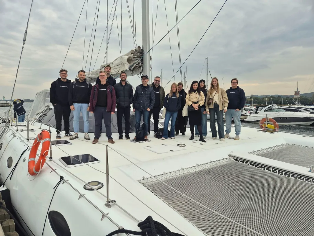 Thirteen colleagues, most in dark Handsontable hoodies, standing on the deck of a white catamaran moored in a Gdynia marina under an overcast sky.