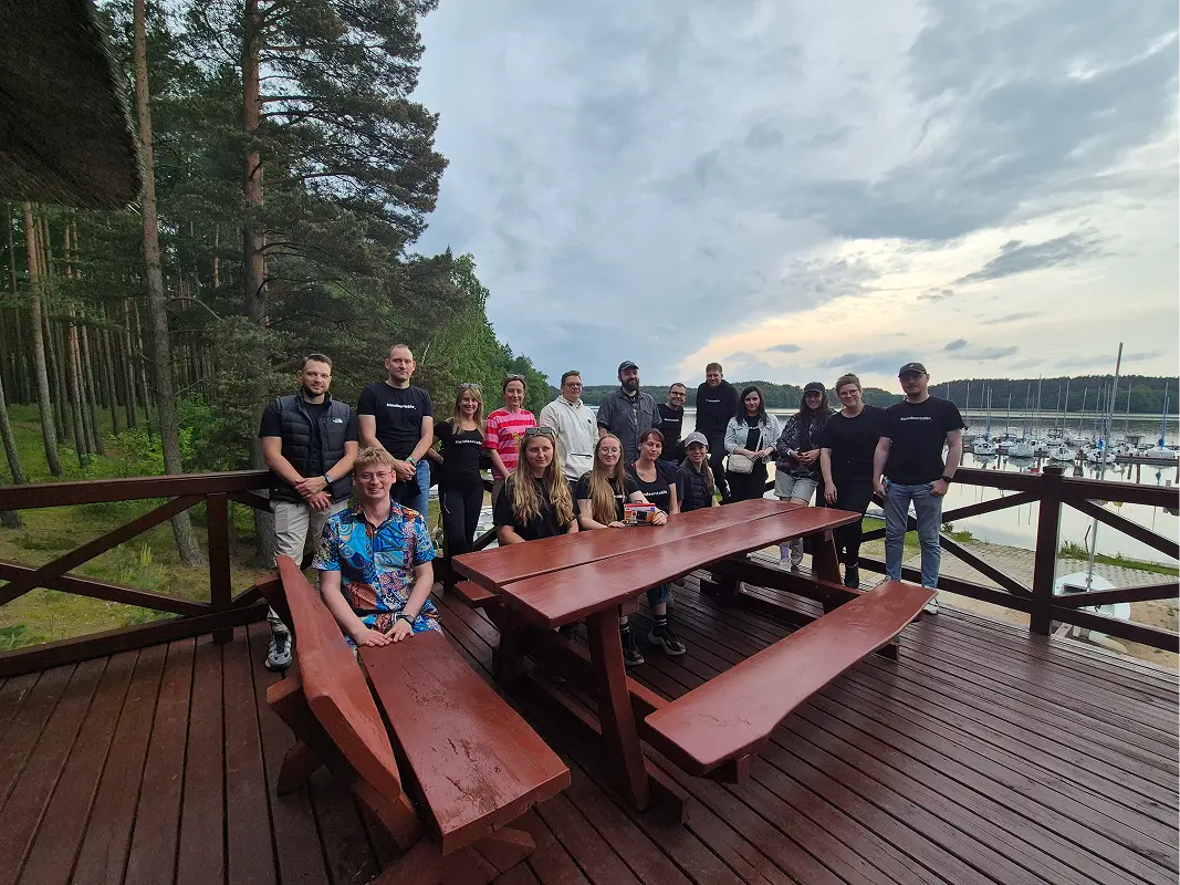 The team gathered around wooden picnic tables on a deck by a lake marina, tall pines on the left.