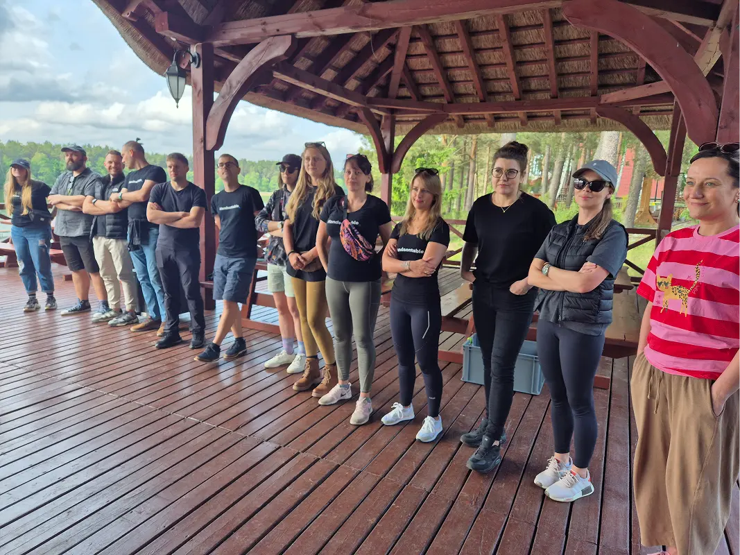 The team lined up along a wooden deck under a thatched gazebo roof, pine trees and water behind them.