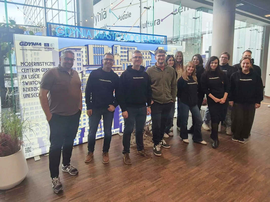 Twelve colleagues, most in dark Handsontable hoodies, standing in front of a lit 'Gdynia Miasto Modernizmu' display in the glass atrium of the Pomeranian Science and Technology Park in Gdynia.