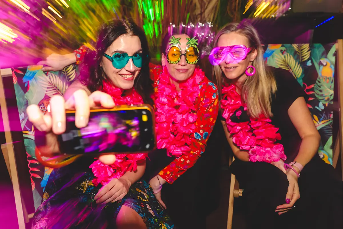 Three colleagues in pink leis and heart-shaped party glasses posing for a phone selfie under neon light.