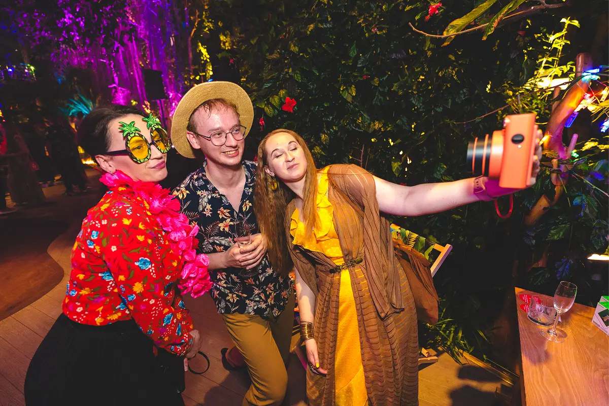 Three colleagues in party glasses and bright shirts taking a selfie among palm leaves at a tropical-themed party.