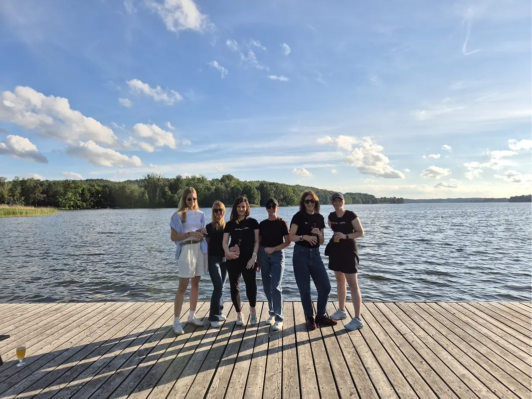 Six colleagues standing on a wooden pier over a lake under a blue sky with white clouds.
