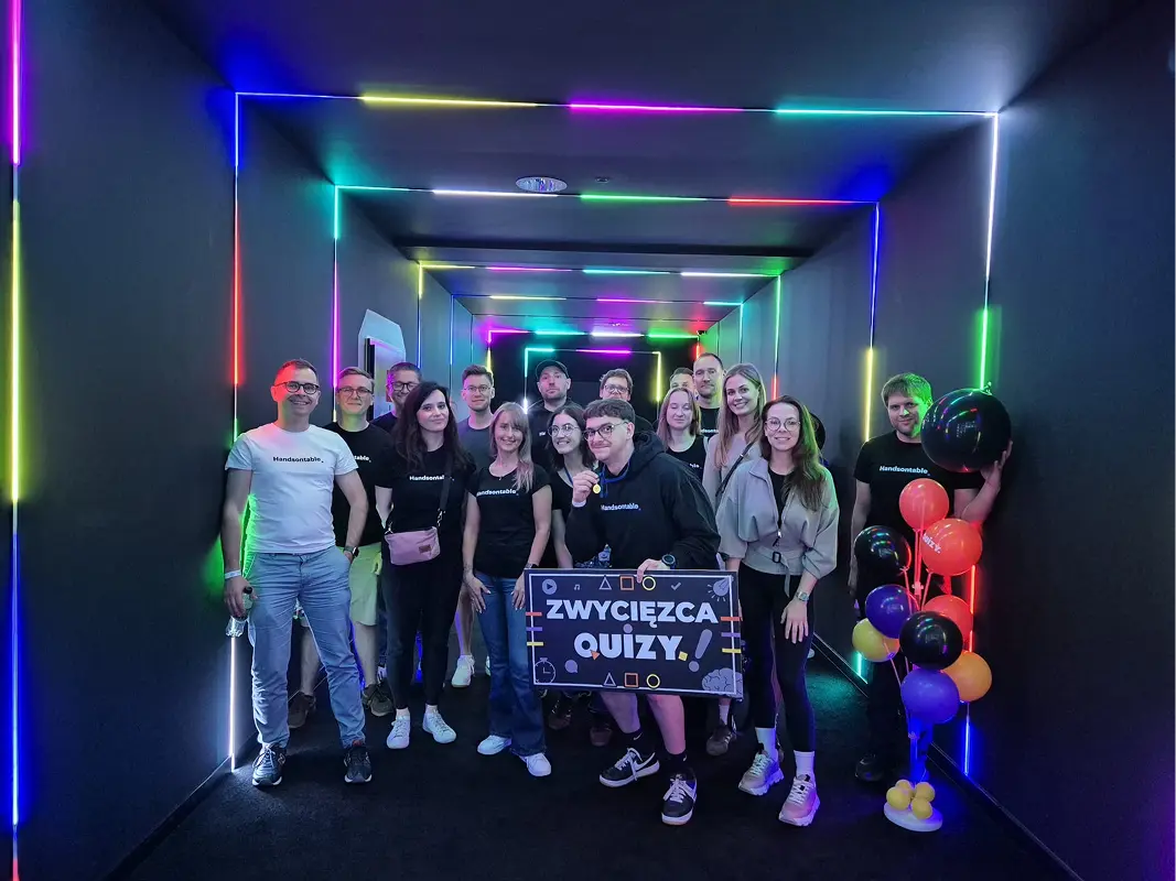 The team in a neon-lit tunnel with balloons, holding a sign that reads Zwycięzca quizu, quiz winner.